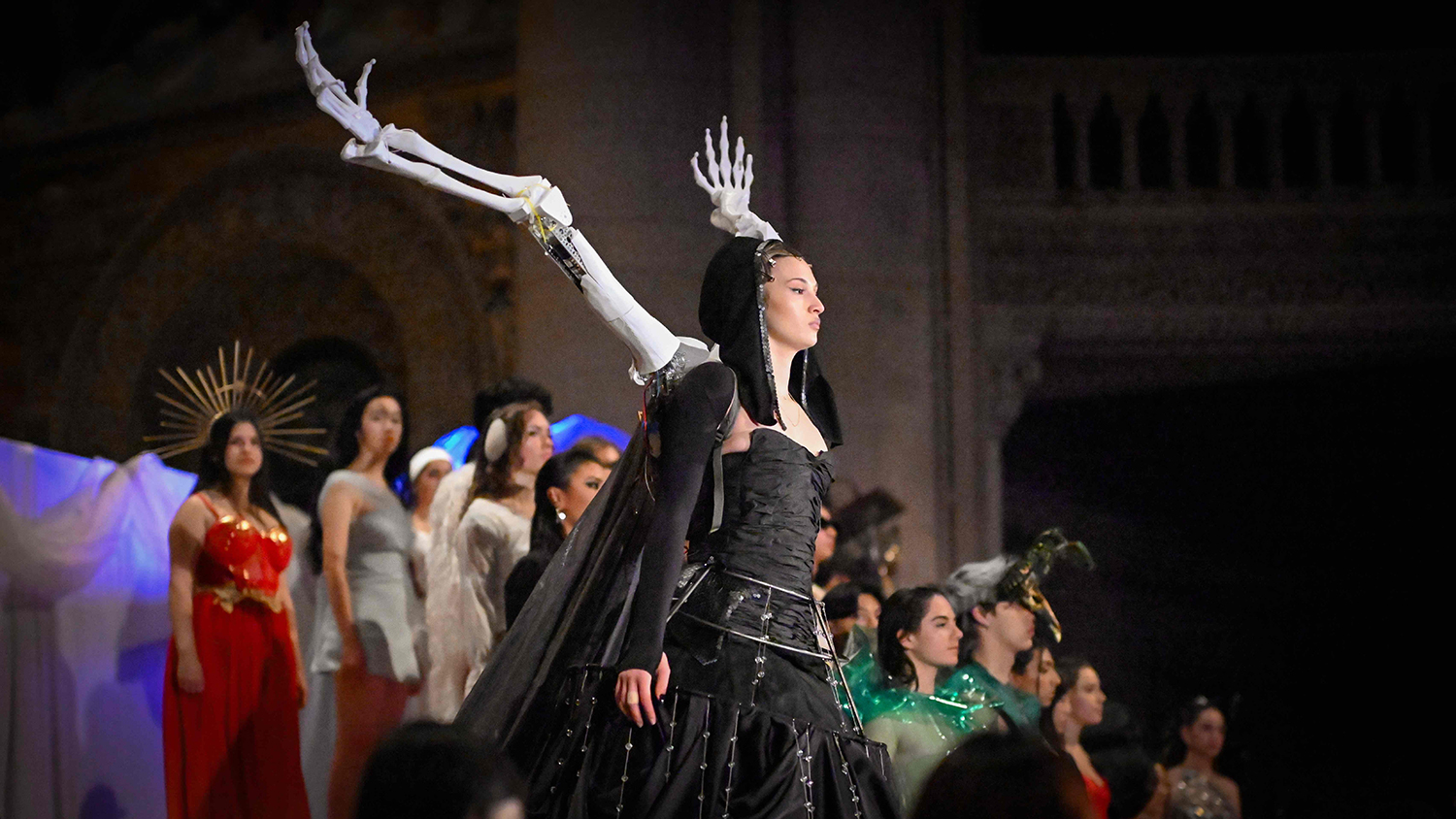 Student model walking down the runway wearing a black dress with two robotic arms coming out the back positioned like wings.