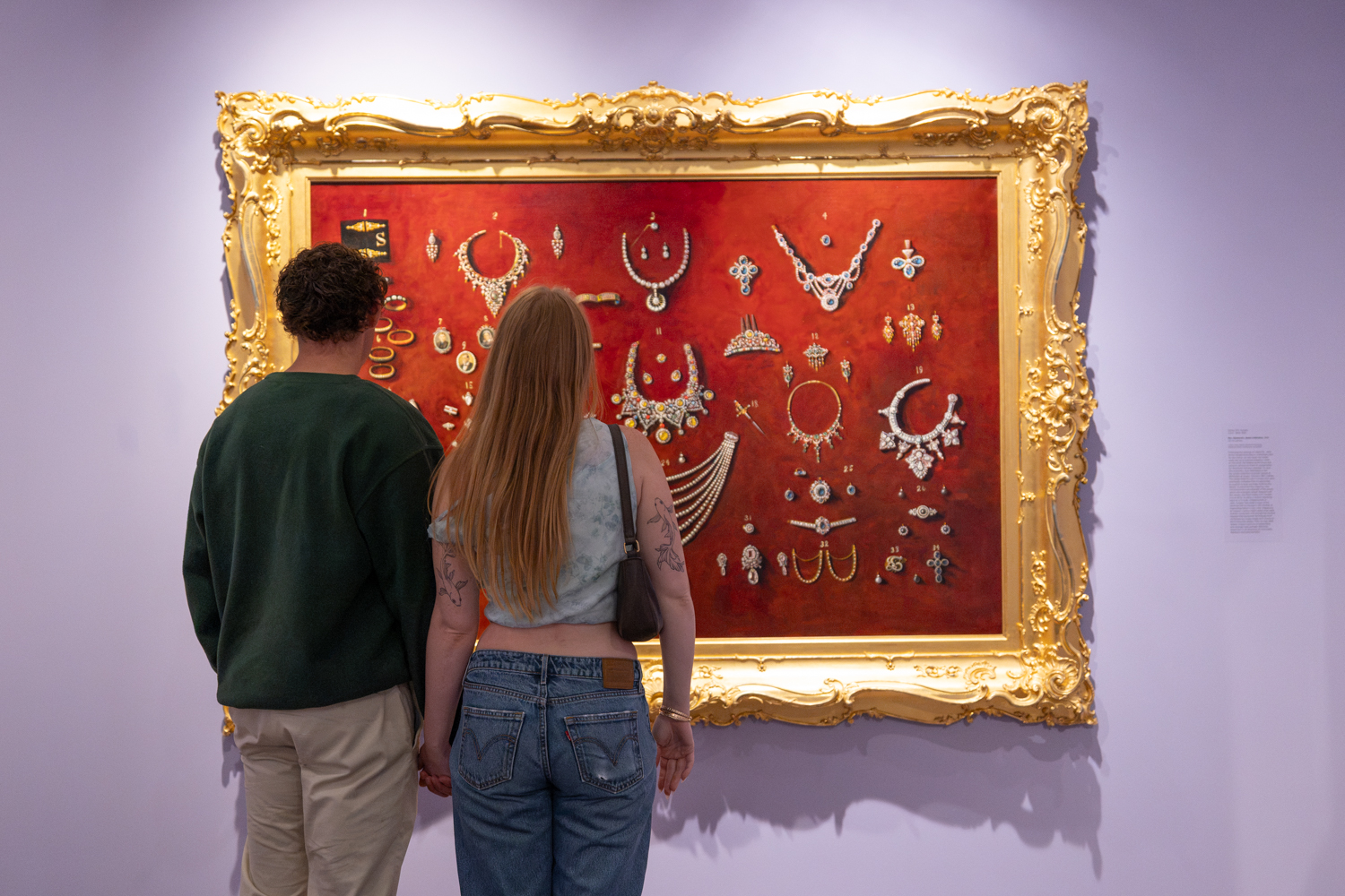 Two visitors admire a large painting of intricate jewelry pieces from Jane Stanford’s collection framed in gold, set against a red background.