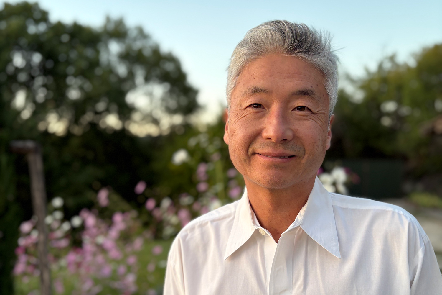 Man with gray hair in a white shirt smiling outdoors with trees and flowers in the background.