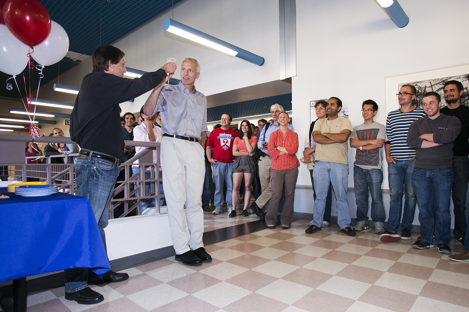 Colleagues gather in a hallway with balloons as Bill Weis raises a glass to toast a smiling Brian Kobilka..