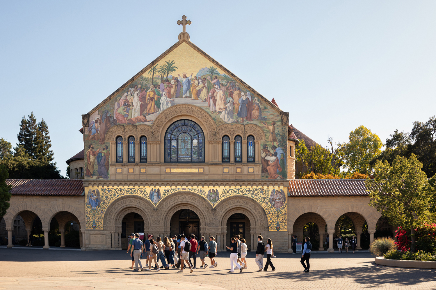 A sunlit Memorial Church with a group of visitors walking across the Quad.