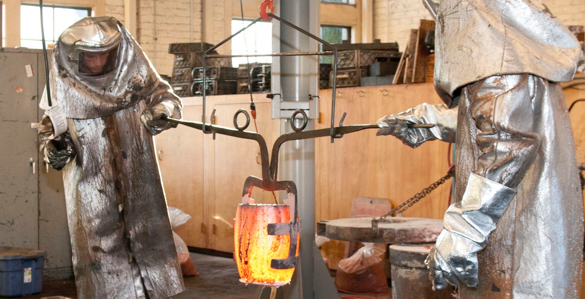 Two people in full-body protective gear moving a container of molten metal