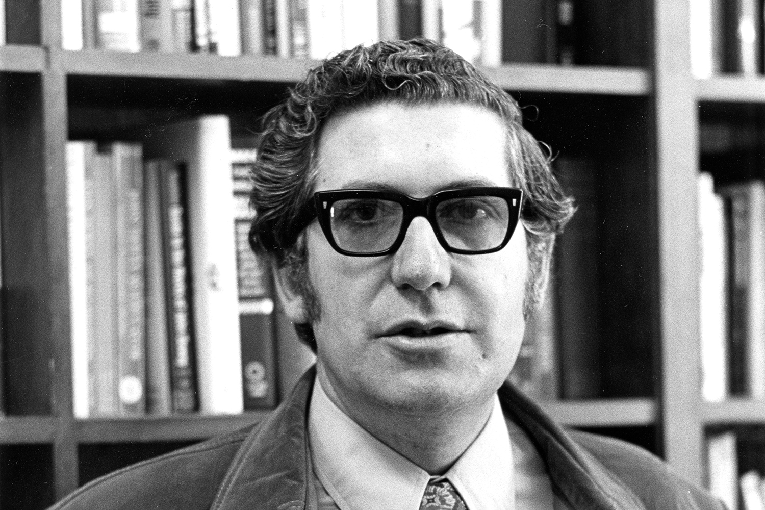 Denis C. Phillips stands in front of bookshelves, wearing a jacket over a button-up shirt and tie, in a black and white portrait.