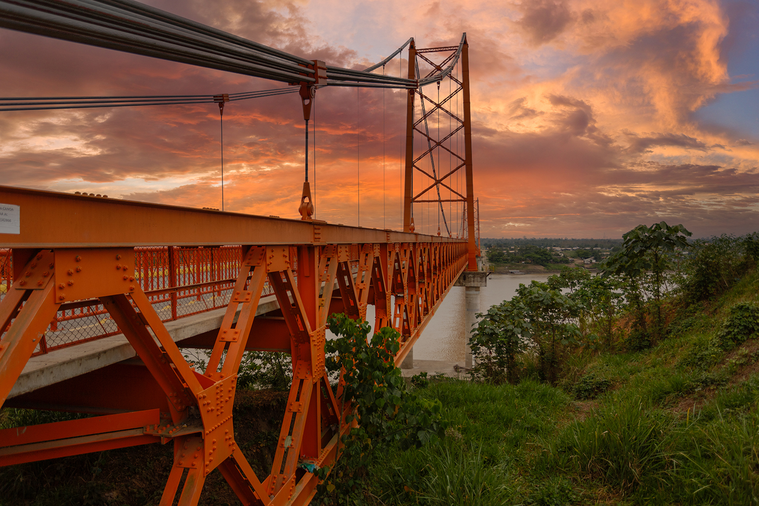 An orange steel suspension bridge stretches over a wide river at sunset beneath a vivid orange and pink sky.