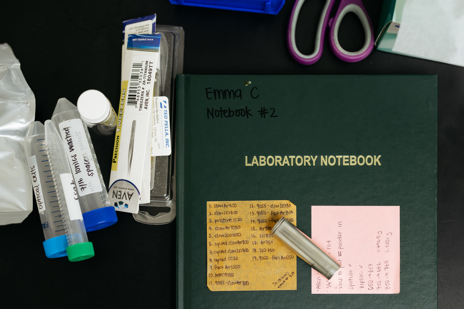 A lab notebook, sample vials, tweezers, and a small metal test cell laid out on a black lab bench.