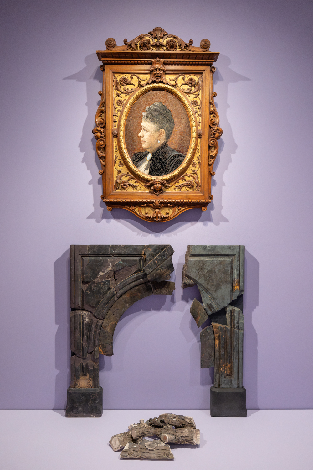 A portrait of a Jane Stanford in ornate framing hangs above two broken architectural elements and scattered fragments, displayed in a museum.