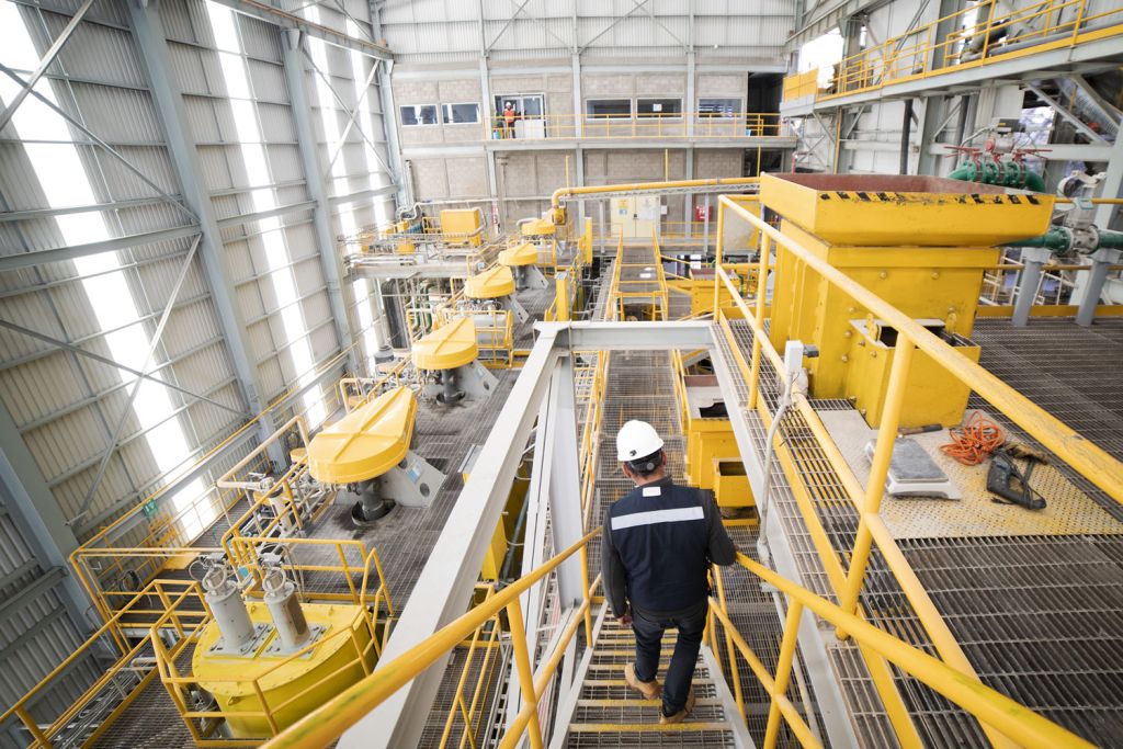 Worker in a hard hat walks down stairs past yellow flotation cells and catwalks inside a large mineral processing plant.
