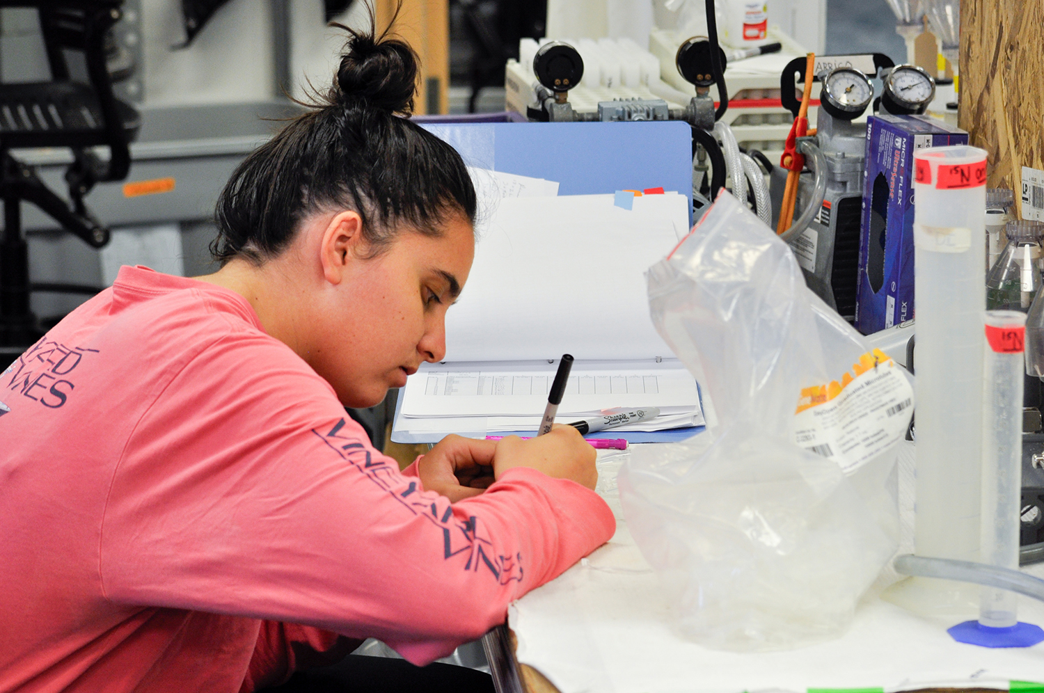 A researcher records data by hand while working at a lab bench filled with sampling equipment.