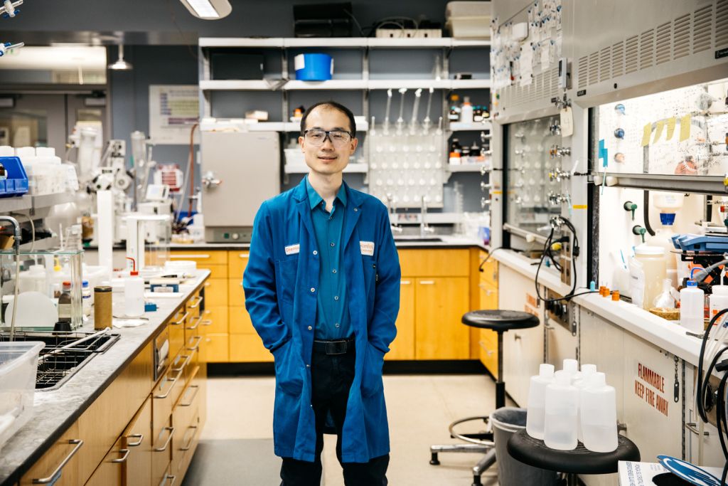 Song Lin in safety glasses and a blue Stanford chemistry lab coat stands in a lab lined with benches and fume hoods.