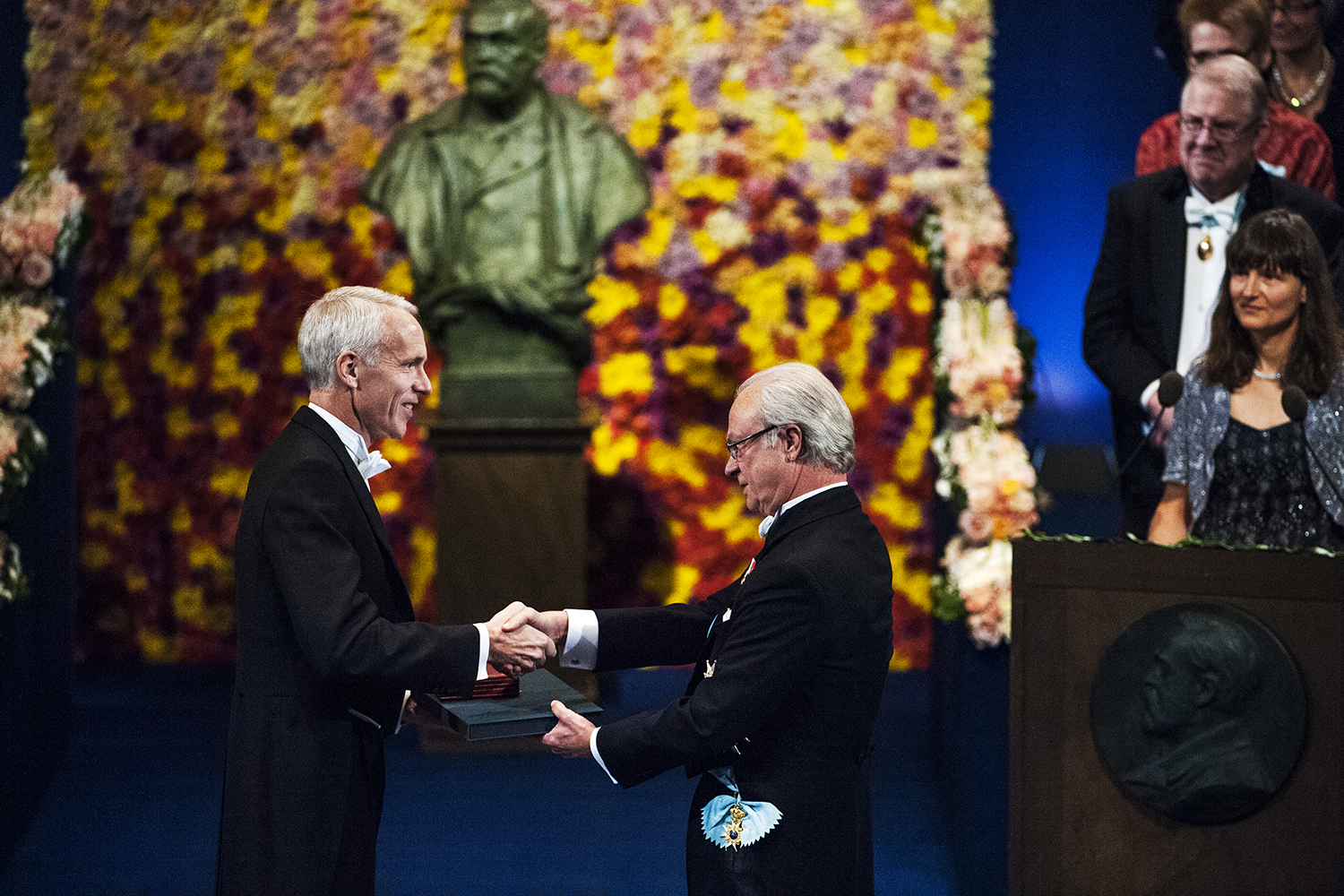 Brian Kobilka in white tie shakes hands with King Carl XVI Gustaf of Sweden as he receives a Nobel award before a flower-framed bust.