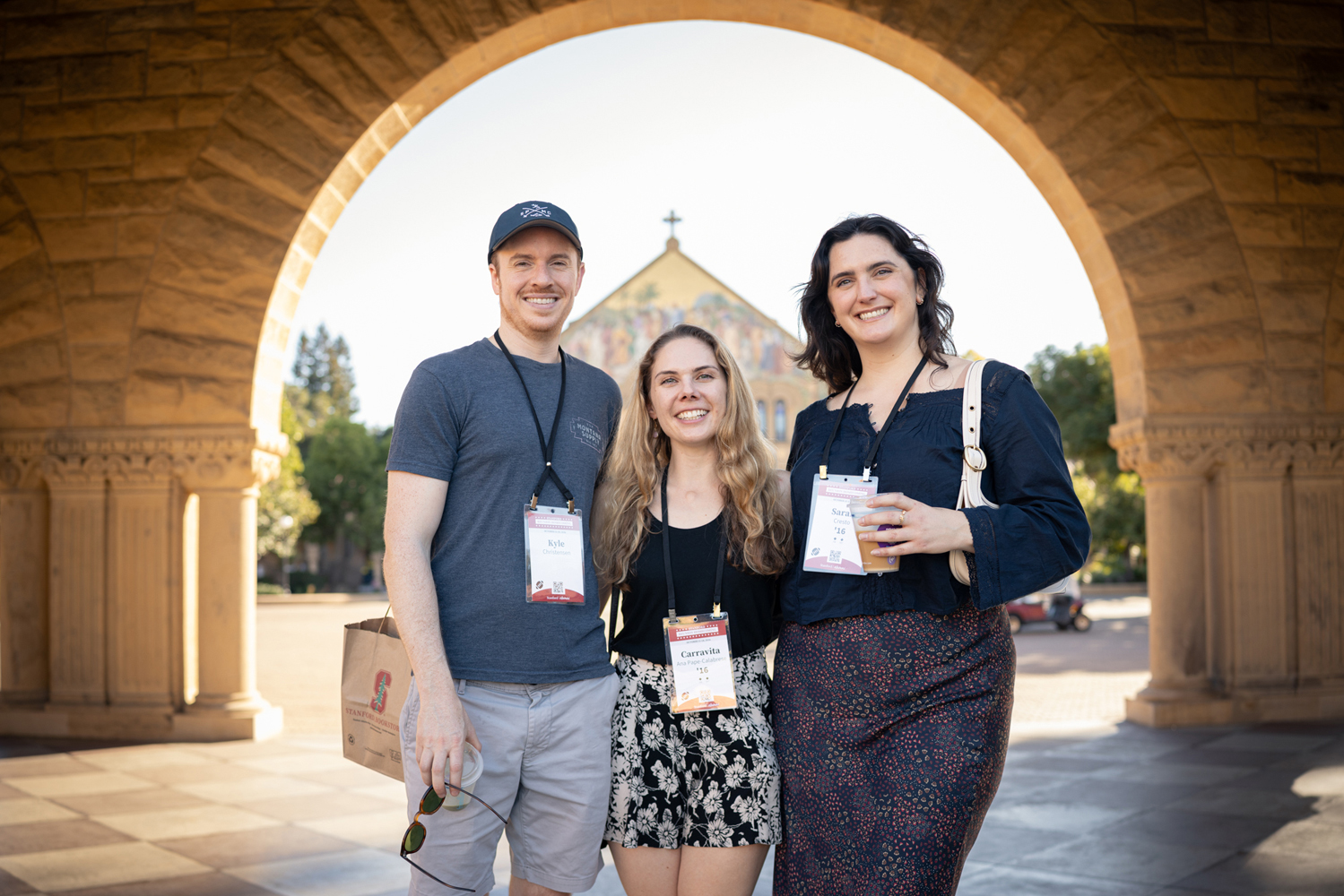 Three smiling alumni pose together under a sandstone arch, with the church facade framed behind them.