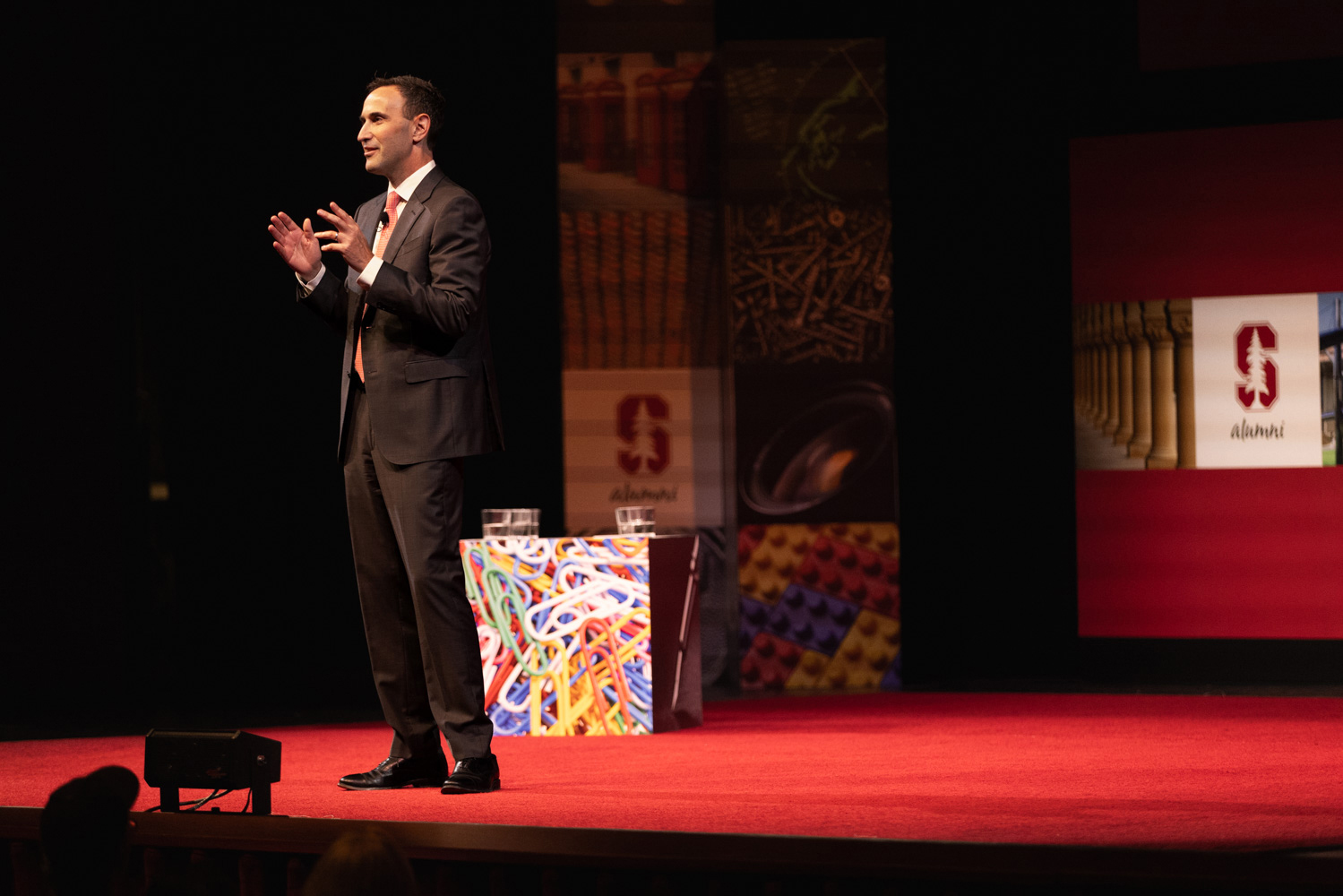 President Jon Levin gestures on a red-carpeted stage with alumni-themed banners and a screen behind him.