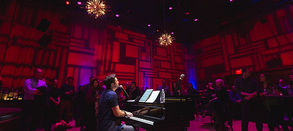 In dramatic red atmospheric lighting, a man plays the piano and sings.