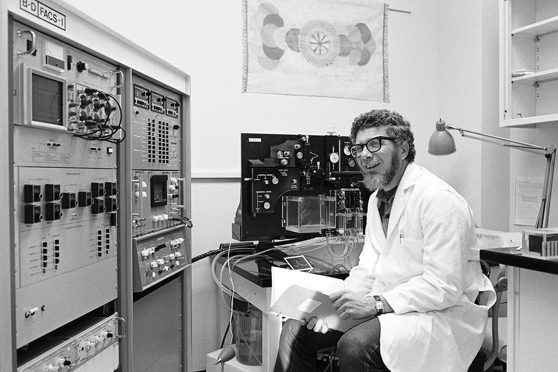 Len Herzenberg in a white lab coat and glasses sitting at a desk with fluorescence-activated cell sorter.