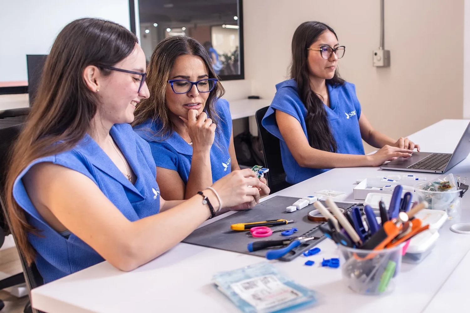 Three individuals in blue tops collaborate at a table with various tools and a laptop, focused on a project.