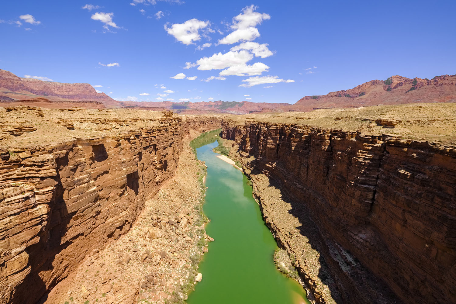Colorado River flowing through a narrow desert canyon with layered red rock walls under a bright blue sky.