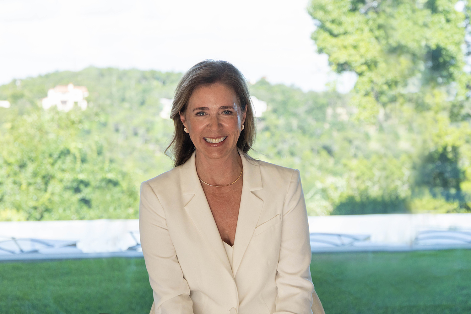 Michelle Landrey Cline in a white blazer smiles while sitting in front of a lush green landscape.