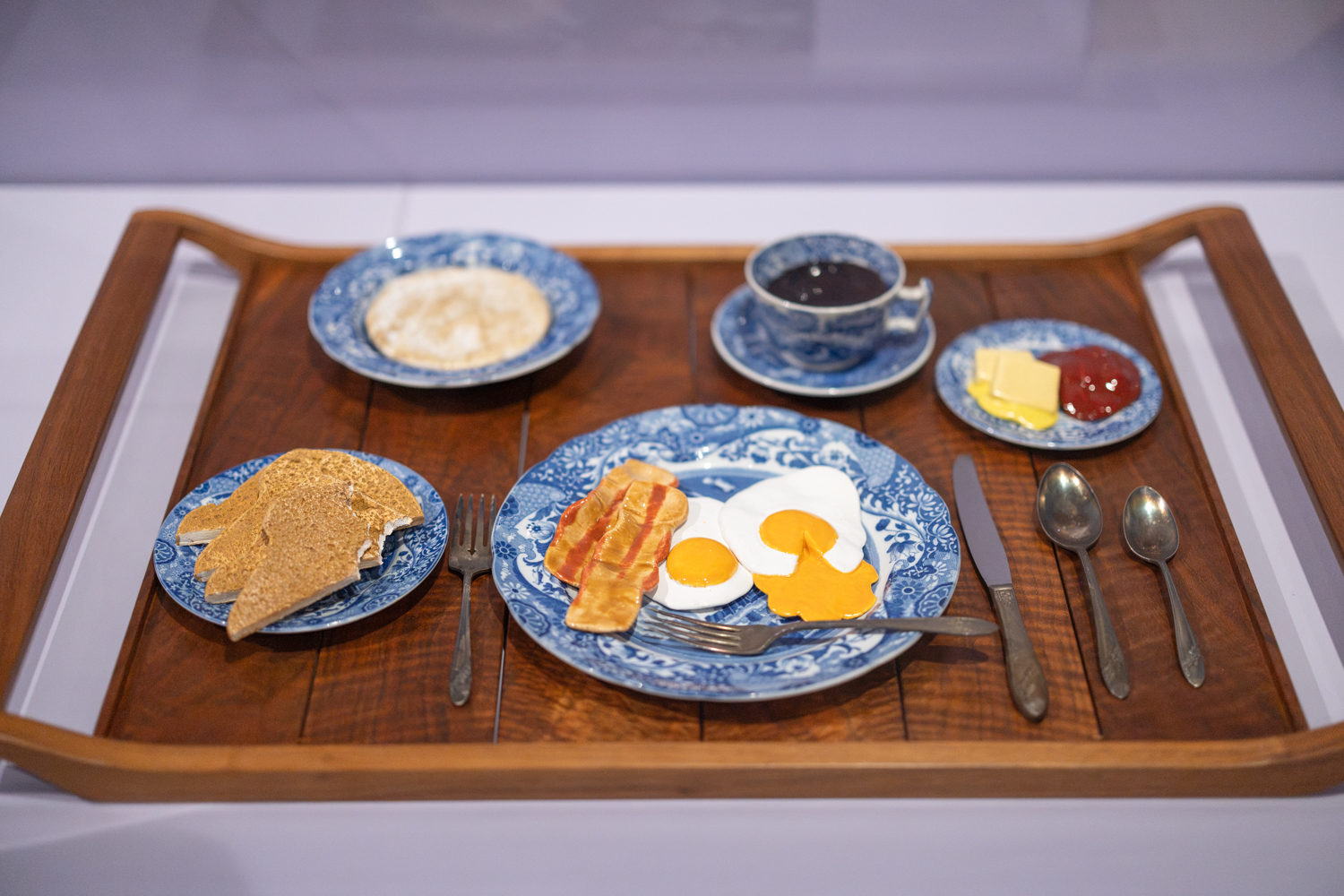 A wooden tray displays a breakfast spread, including scrambled eggs, bacon, toast, coffee, and condiments, all served on blue patterned dishes.