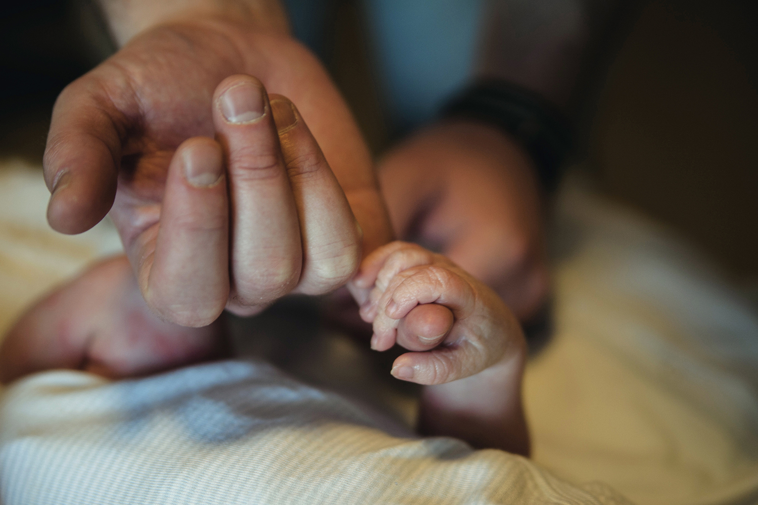 A newborn baby grips an adult's finger while lying wrapped in a blanket.