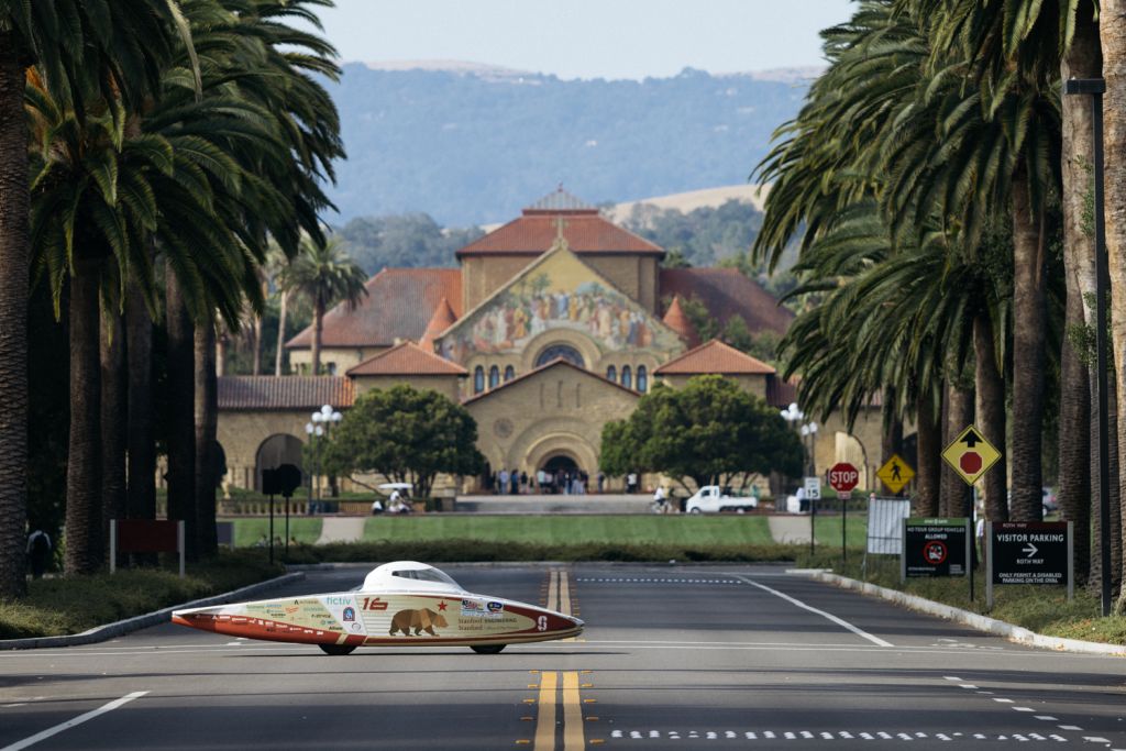Stanford's solar-powered race car on an empty campus road, framed by tall palm trees with Memorial Church behind it.