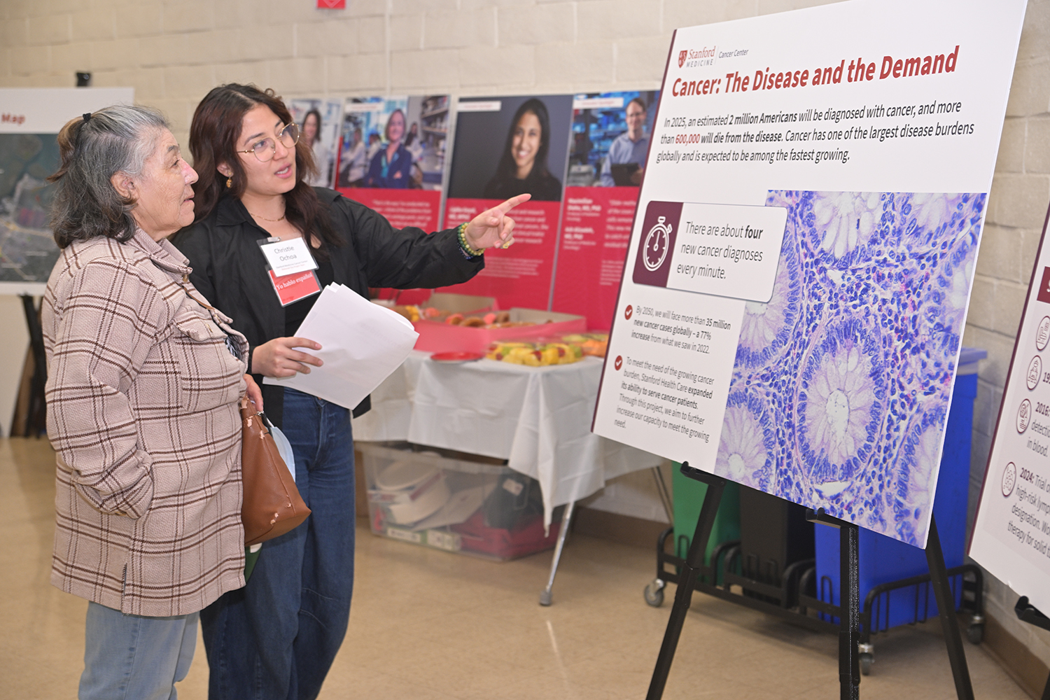 A staff member wearing a name badge explains a cancer research poster's key statistics to a visitor at a health fair.
