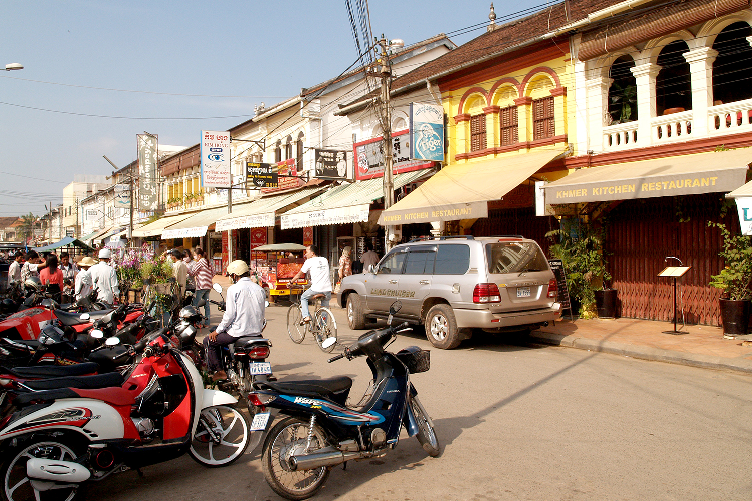 A busy street scene in a market with shops and restaurants, featuring parked motorcycles and people walking and cycling along the road.
