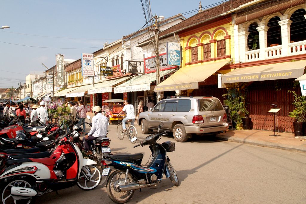 A busy street scene in a market with shops and restaurants, featuring parked motorcycles and people walking and cycling along the road.