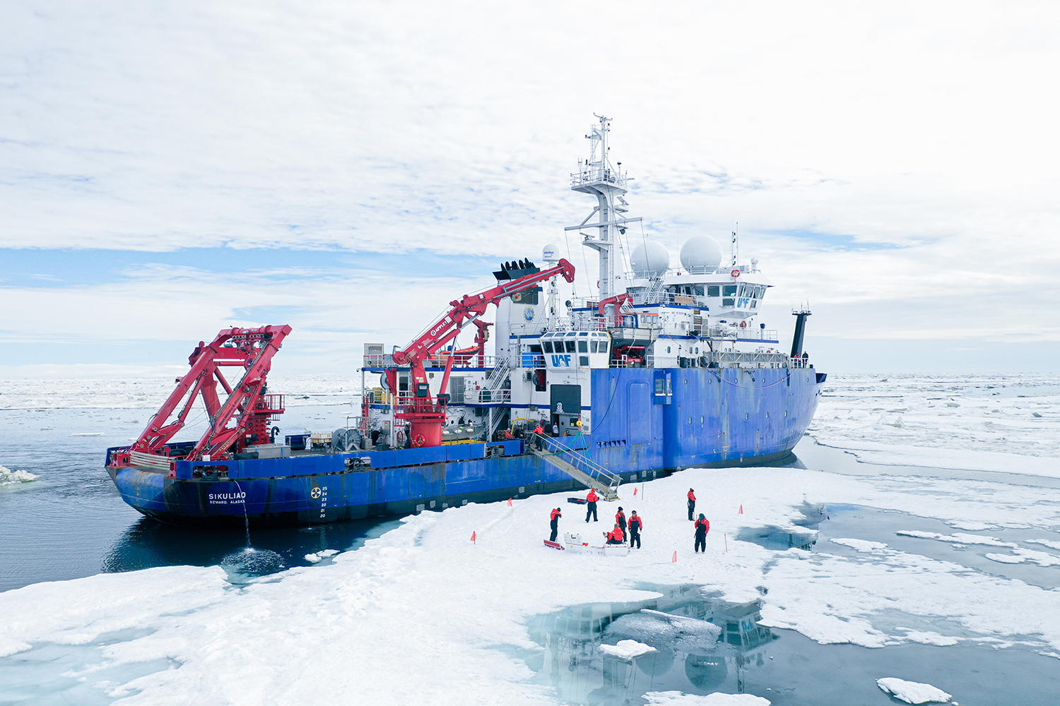 A research vessel sits docked against Arctic sea ice as scientists collect samples nearby.