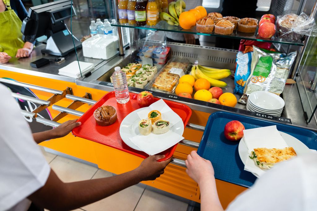 Two students slide lunch trays with wraps, muffins, apples and flatbread along a cafeteria counter stocked with fruit and snacks.