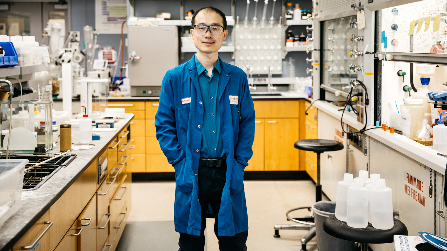 Song Lin in safety glasses and a blue Stanford chemistry lab coat stands in a lab lined with benches and fume hoods.