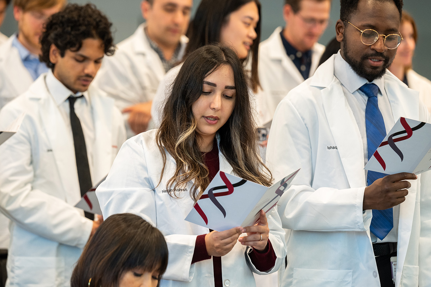 Graduate students in white coats stand in rows holding printed programs with a DNA helix design, reading aloud at a coat ceremony.