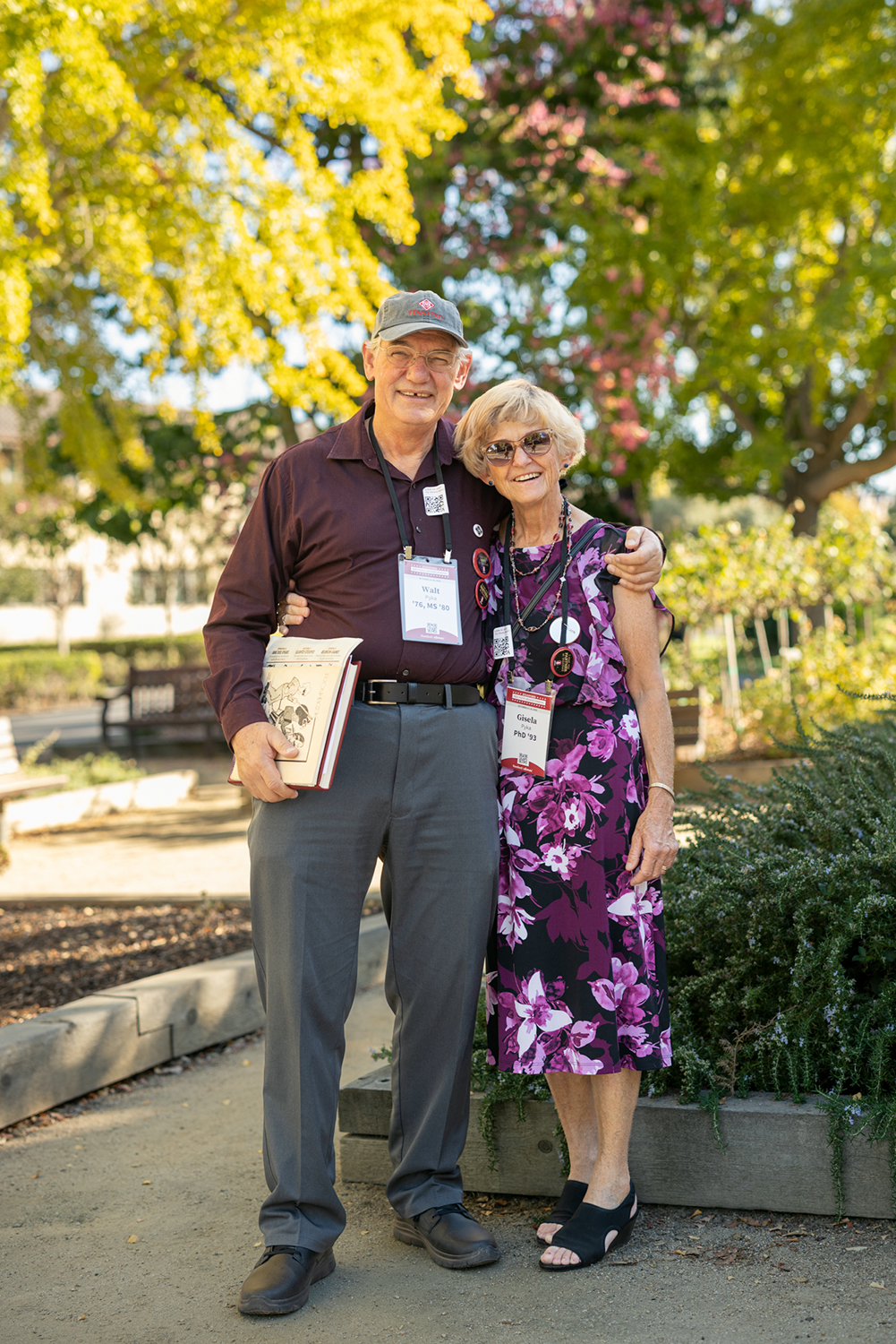 Smiling couple stands arm in arm on a garden path on the Stanford campus, the man holding a yearbook.