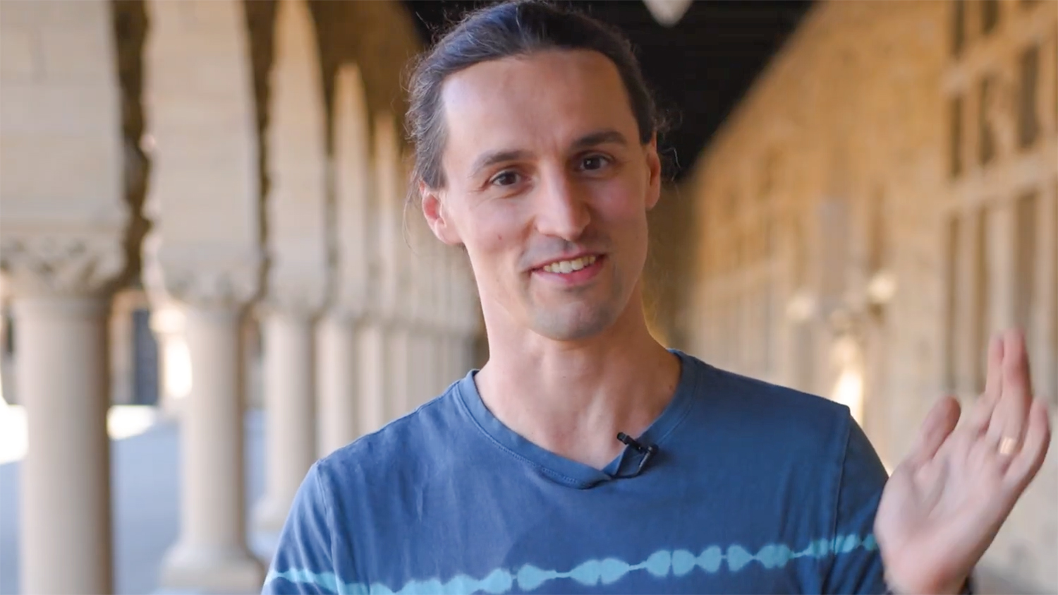 Ettore Biondi, wearing a clip-on microphone, waves gestures with his hands while talking in a sunlit sandstone arcade.