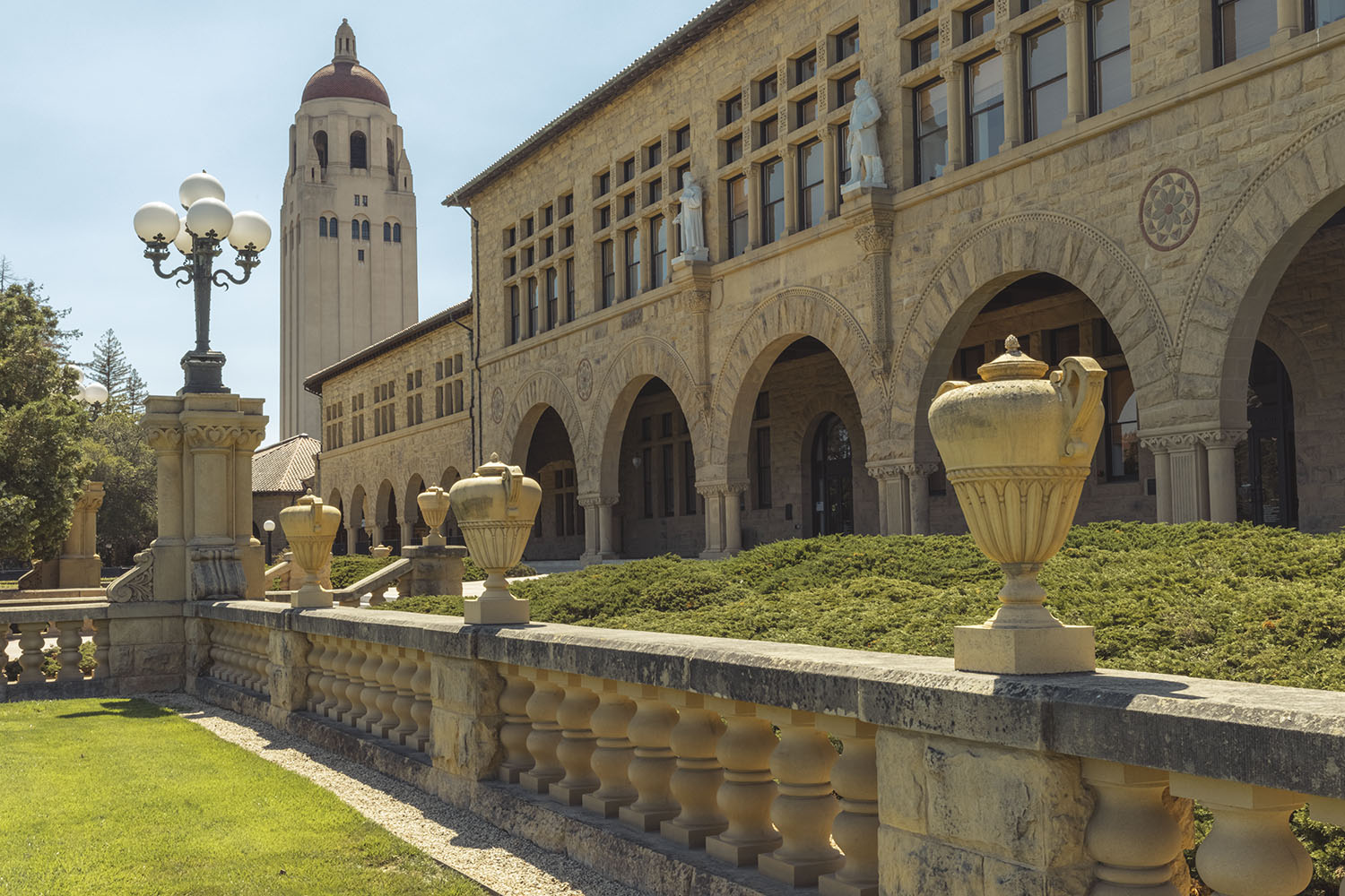 The stately Wallenberg Hall is seen behind a low stone fence and some hedges. In back, Hoover Tower stands.