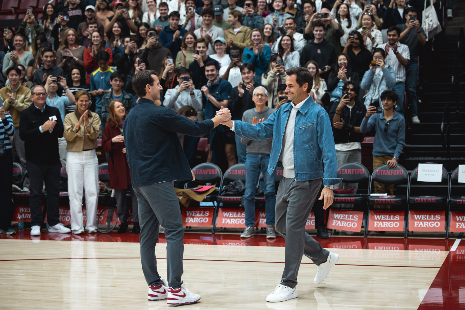 Roger Federer and Nico Godsick shake hands on a basketball court while a cheering crowd watches from the stands.