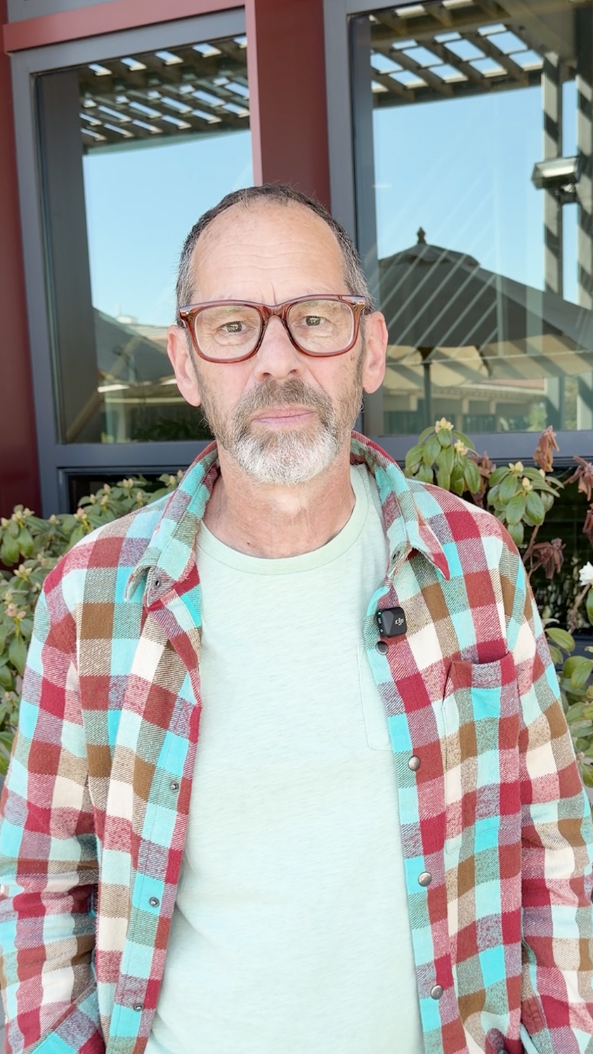 Dan Jurafsky stands near a building, wearing a colorful plaid shirt over a light green t-shirt,