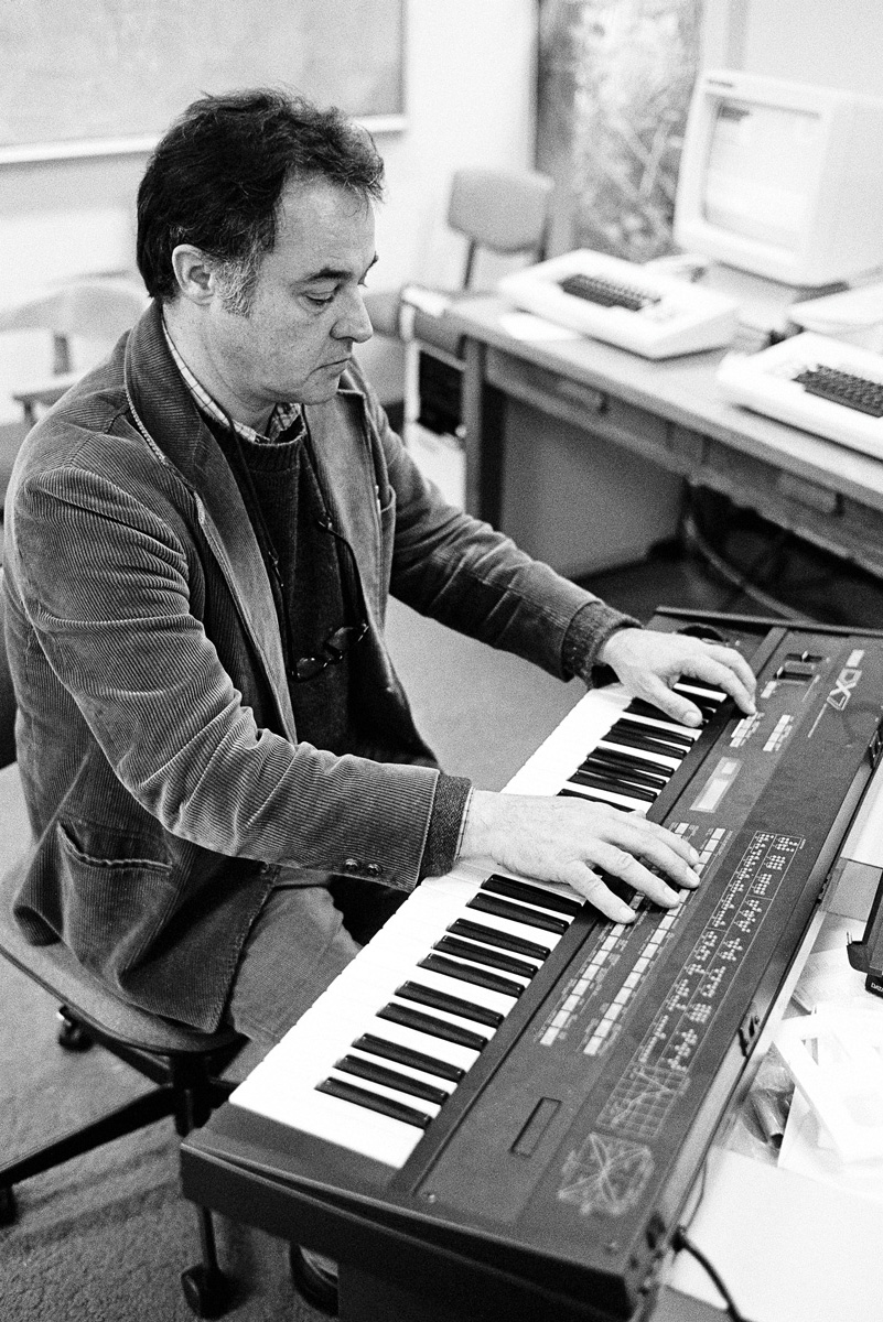 John Chowning in a corduroy jacket playing an electronic keyboard in a room with computers.