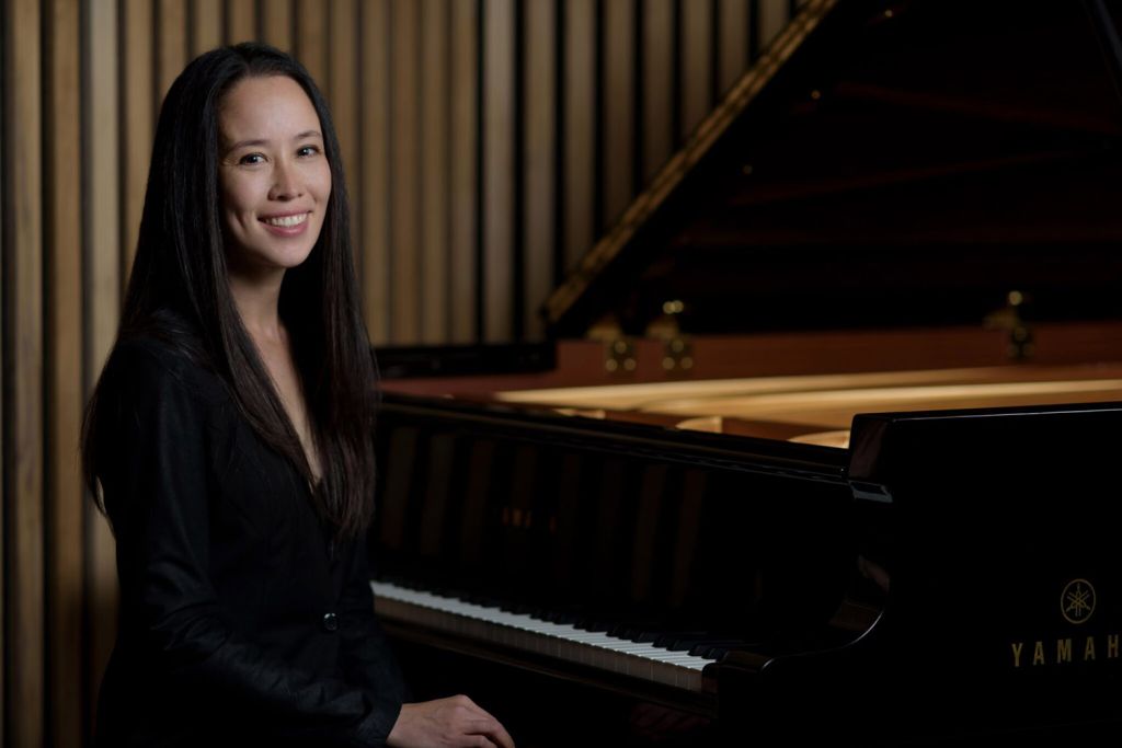 Pianist smiles warmly while sitting beside a grand Yamaha piano in a wooden-paneled room.
