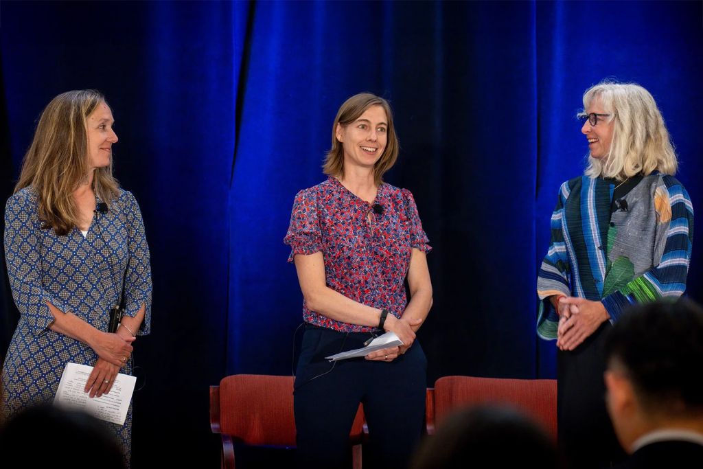 Three women engage in discussion on stage at an event, with microphones and seated chairs visible in the background.
