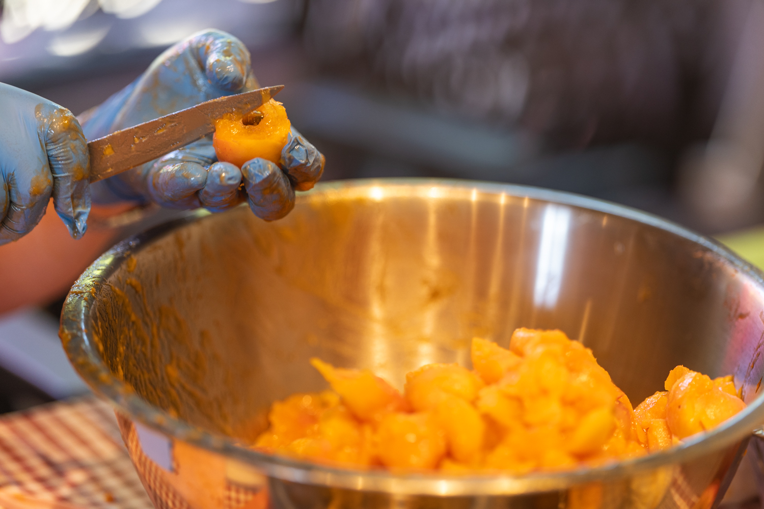 Gloved hand using a corer to remove apricot pits over a metal bowl.