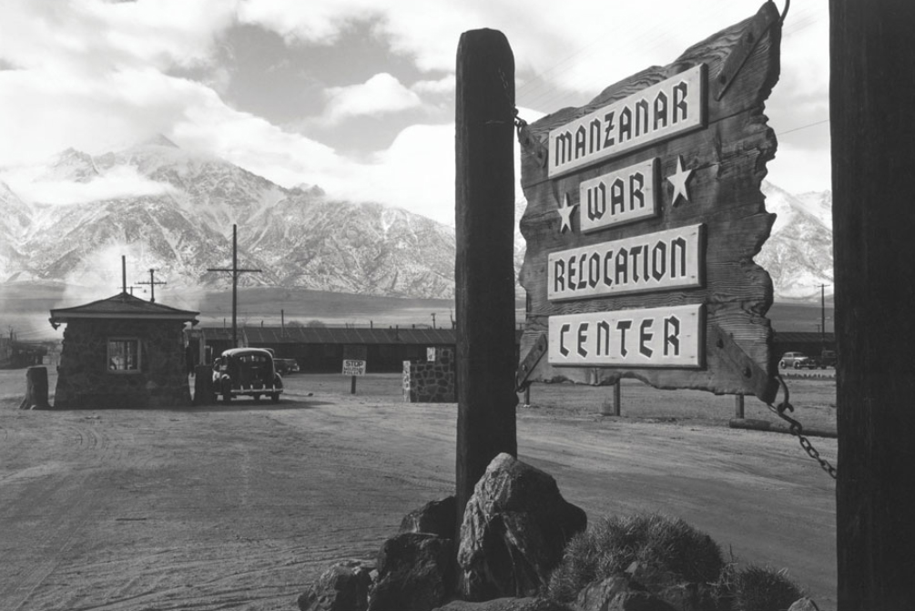 Black-and-white photo of the entrance sign to Manzanar War Relocation Center, with a guard post and snow-capped mountains in the background.