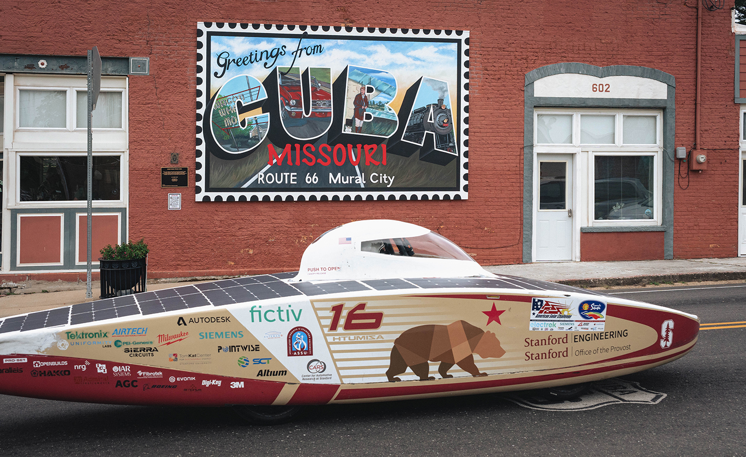 Stanford's solar car parked along a street below a large postcard-style mural welcoming visitors to Cuba, Missouri.