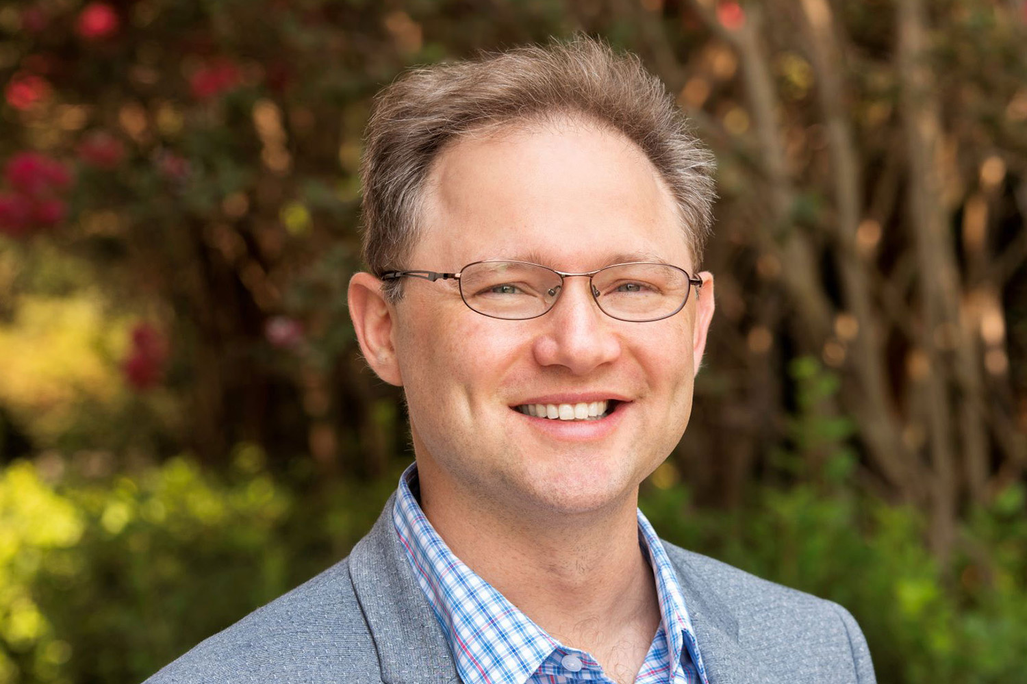 A smiling man with glasses wears a gray blazer over a checked shirt, with green foliage and flowers in the background.