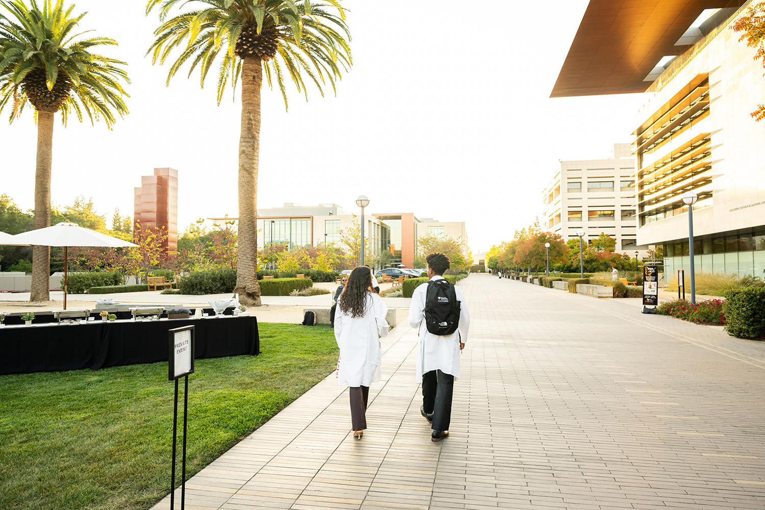 Two people in white lab coats walk away from the camera along a brick campus path lined with palm trees and modern buildings at sunset.