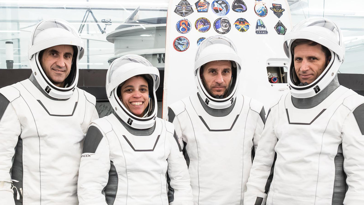 Four people in white spacesuits and helmets stand side by side in front of a capsule panel covered in mission patches.