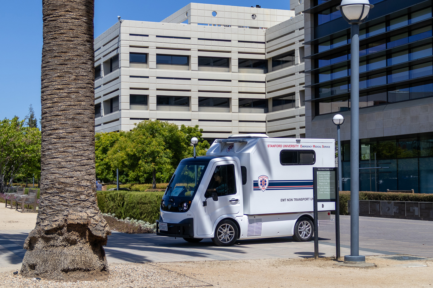 Small white emergency medical service vehicle parked on a campus walkway between a palm trunk and modern buildings.