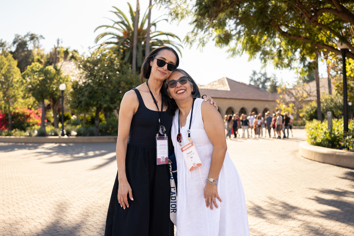 Two women smile for a photo in a sunny plaza with palm trees and a crowd gathered in the distance.