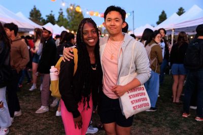 Two friends smiling in the foreground, festival booths in the background.