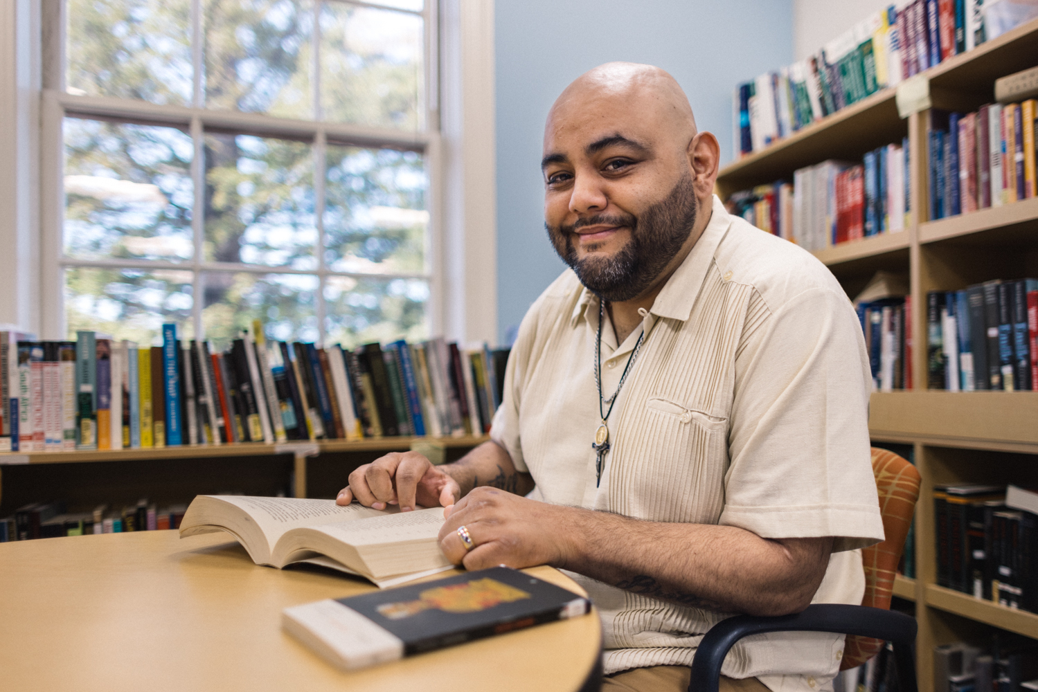 Christian Sanchez, open book in hand, smiles in a brightly-lit library.