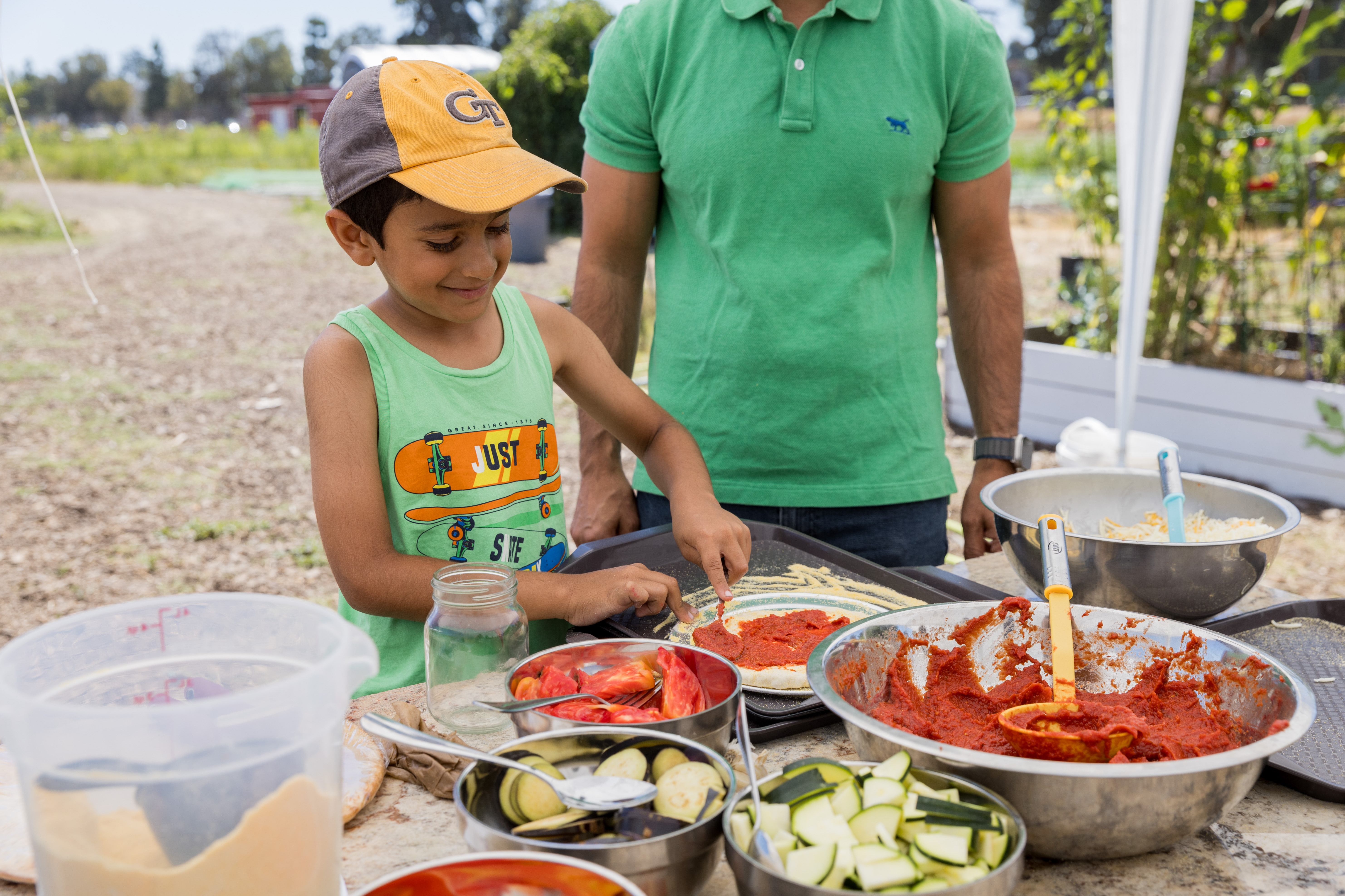 A little boy in a green tank top and baseball cap grins as he spreads sauce on a round pizza dough on a table surrounded by bowls of sliced ​​vegetables and other fresh ingredients. 